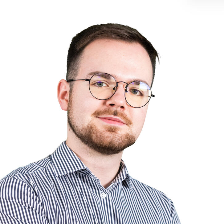 Zdjęcie portretowe. Mężczyzna z brązowymi włosami w okularach i zarostem. Lekko uśmiechnięty.